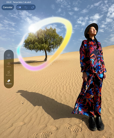 Parte da frente do iPad Pro preto-espacial na vertical, bordas arredondadas, tela com a imagem de uma mulher de vestido colorido parada em uma paisagem desértica com uma única árvore ao fundo contornada por um degradê arco-íris luminoso, mostrando a capacidade da Apple Intelligence de apagar objetos em segundo plano