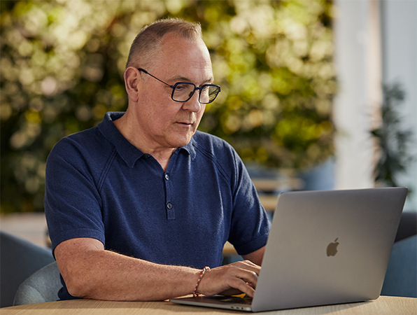 Az Apple szoftverekkel és szolgáltatásokkal foglalkozó alkalmazottja a szabadban dolgozik a MacBookján, n?vényekkel a háttérben.