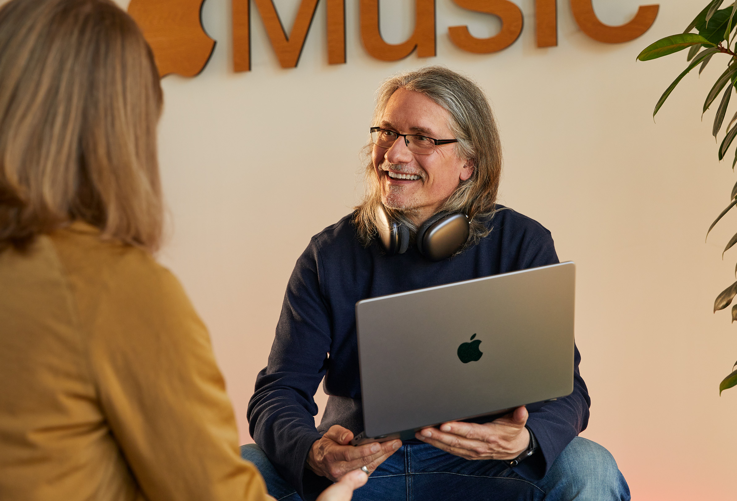 Két Apple-alkalmazott beszélget, az egyik MacBookot tart a kezében és egy Apple?Music logót ábrázoló fal el?tt ül.