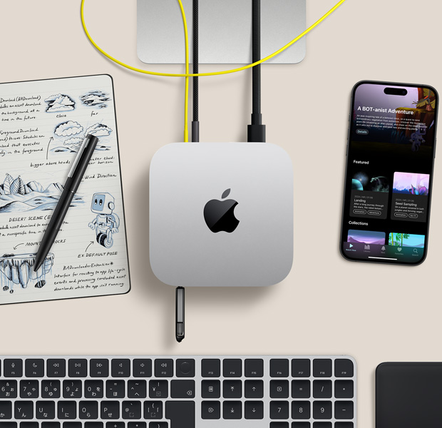 デスク上に置かれたシルバーのMac miniを上から見た図。コードとフラッシュドライブがポートにつながれている。ワイヤレスキーボード、トラックパッド、iPhone、ノートが一緒に配置されている