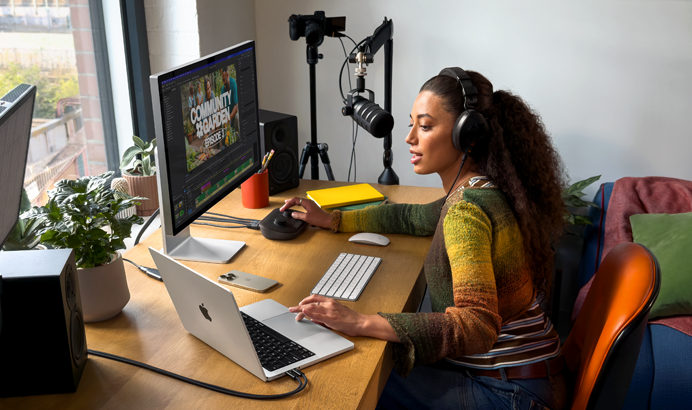 Sieviete savās mājās pie rakstāmgalda strādā ar MacBook Pro, kas savienots ar diviem ārējiem displejiem, – vi?a ir uzlikusi austi?as un runā mikrofonā