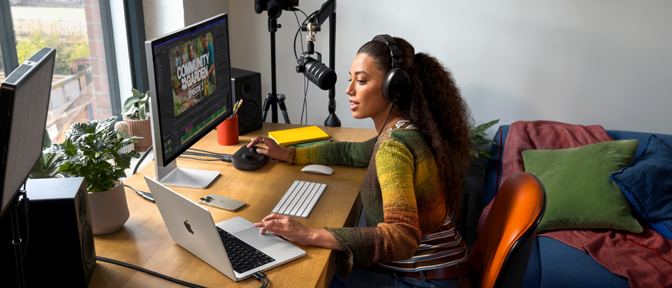 O femeie acas? lucreaz? la birou cu un MacBook Pro conectat la dou? monitoare externe, purtand c??ti ?i vorbind ?ntr-un microfon
