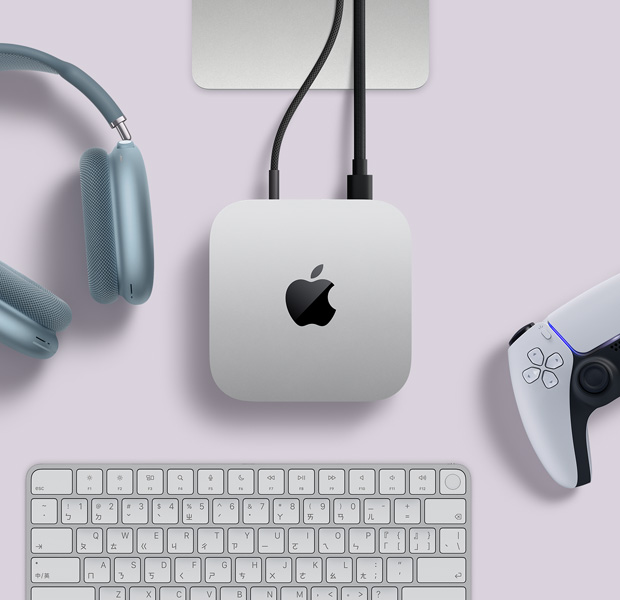 銀色 Mac mini 的俯視圖，兩條連接線插入背面連接埠，旁邊放著藍(lán)色 AirPods Max、銀色巧控鍵盤和無線遊戲控制器