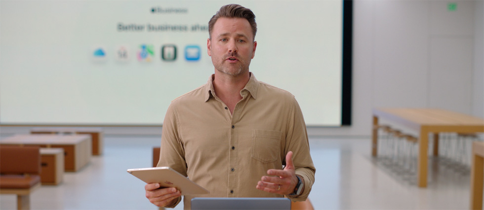 Présentateur parlant avec un iPad à la main, debout devant un MacBook posé sur une table, tandis qu’un grand écran derrière lui affiche les ic?nes de plusieurs apps professionnelles